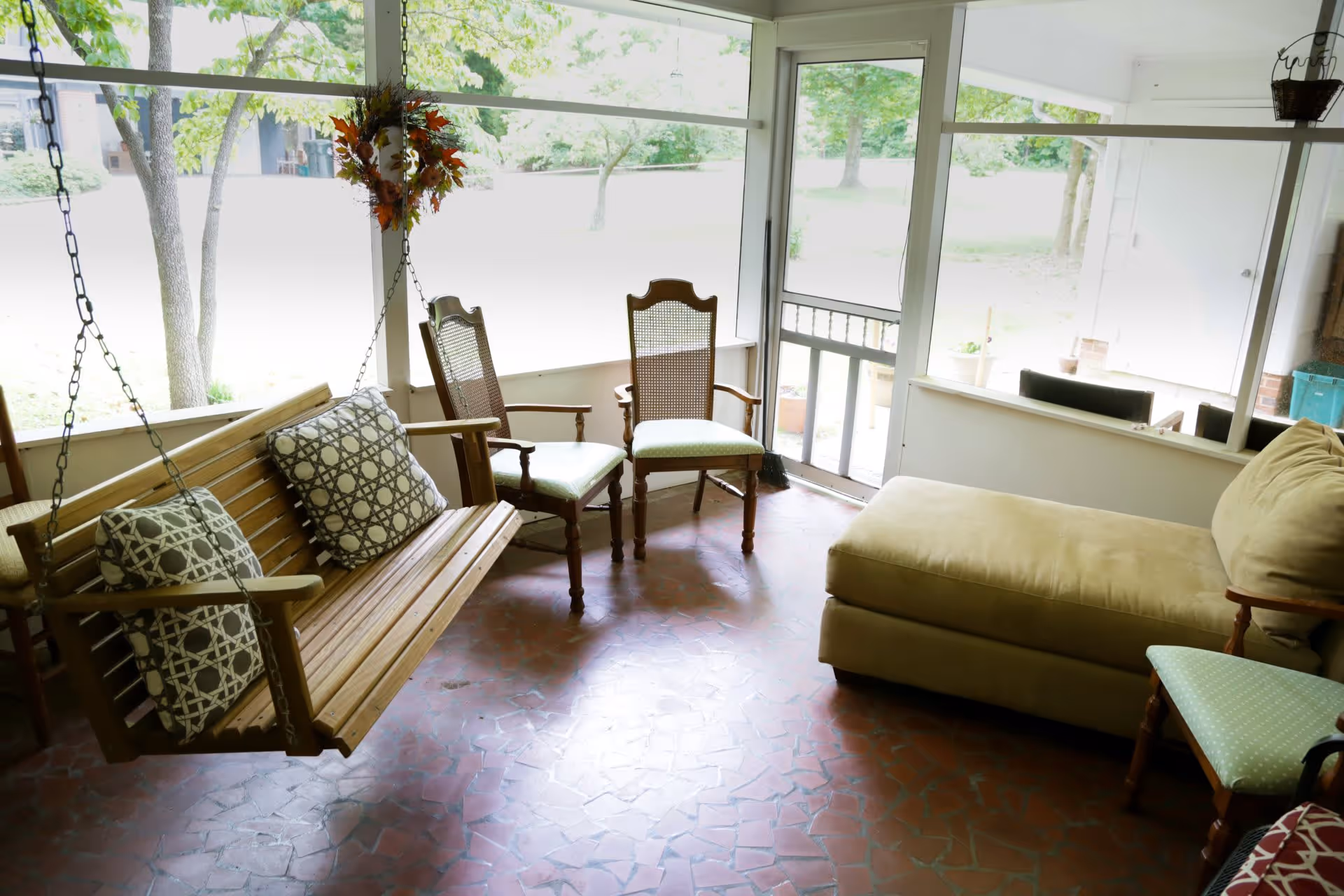 Sunroom with a wooden hanging swing, several chairs, and an upholstered chaise overlooking a grassy yard.