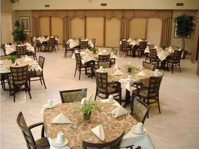 A spacious dining room with multiple round tables covered with patterned tablecloths and white napkins folded into triangles. Each table has a small green plant centerpiece and is surrounded by wooden chairs with cushioned seats. The room has large windows with beige blinds and decorative valances, and there are a few potted plants along the walls.