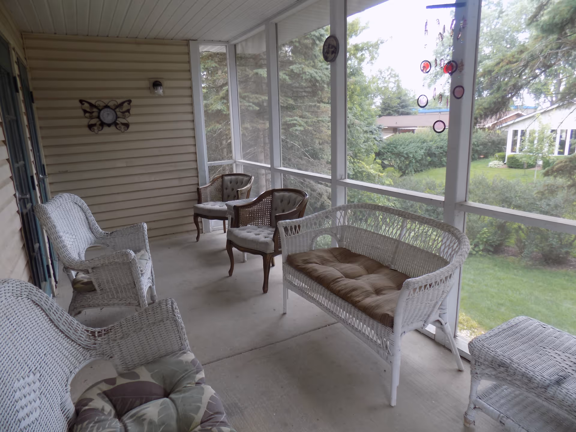 A screened-in porch with several cushioned wicker chairs and a wicker loveseat. The porch overlooks a green yard with trees and bushes, and there is a decorative butterfly clock on the wall.