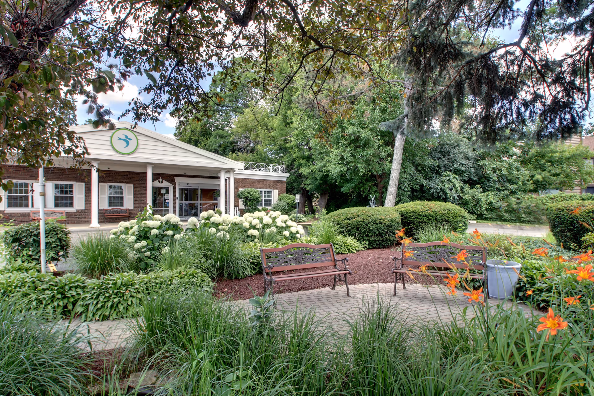Outdoor garden area at BRIA of Geneva featuring two metal benches on a paved path surrounded by green bushes, white flowers, and orange lilies. The building entrance with white columns and a circular logo is visible in the background, partially shaded by tree branches.
