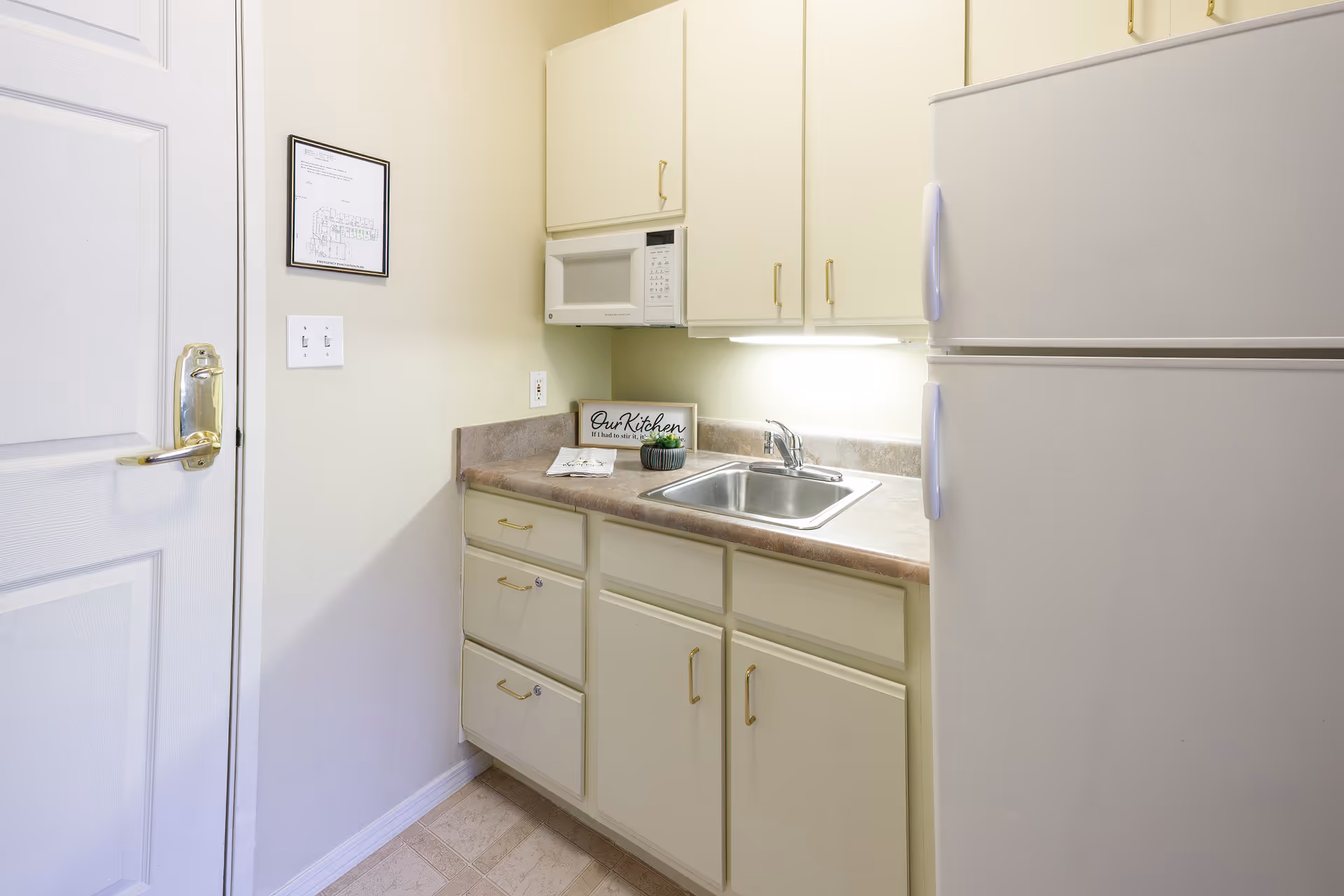 Small kitchen area with white cabinets, a countertop with a sink, a white microwave mounted above the counter, and a white refrigerator. There is a framed sign on the counter that reads 'Our Kitchen' along with a small decorative plant. A door with a gold handle is visible on the left side of the image.