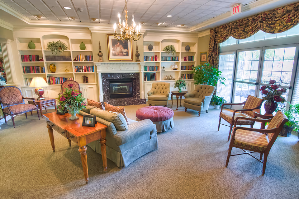 A cozy living room area in a senior living facility with a fireplace surrounded by built-in bookshelves filled with books and decorative items. The room features a chandelier, a sofa with cushions, two armchairs, a round ottoman, wooden chairs, side tables with plants and flowers, and large windows with curtains letting in natural light.