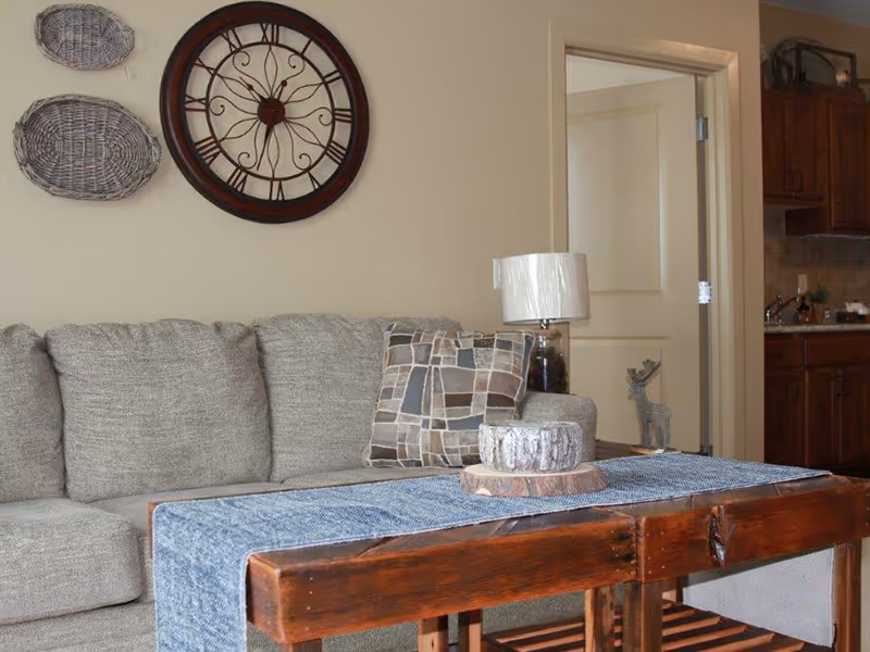 Cozy living room with a gray sofa, wooden coffee table with a runner and decorative centerpiece, a large wall clock and woven baskets, and a view into a kitchen doorway.