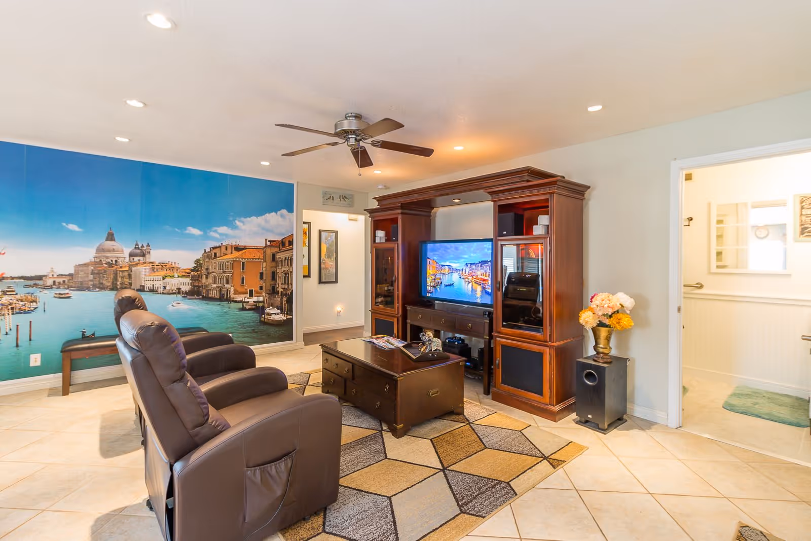 A cozy living room with two brown recliner chairs facing a wooden entertainment center with a flat-screen TV displaying a scenic canal view. The room features a ceiling fan, a geometric patterned rug, a coffee table, and a large wall mural of a waterfront cityscape. There is a doorway leading to a bathroom with white walls and a green bath mat.