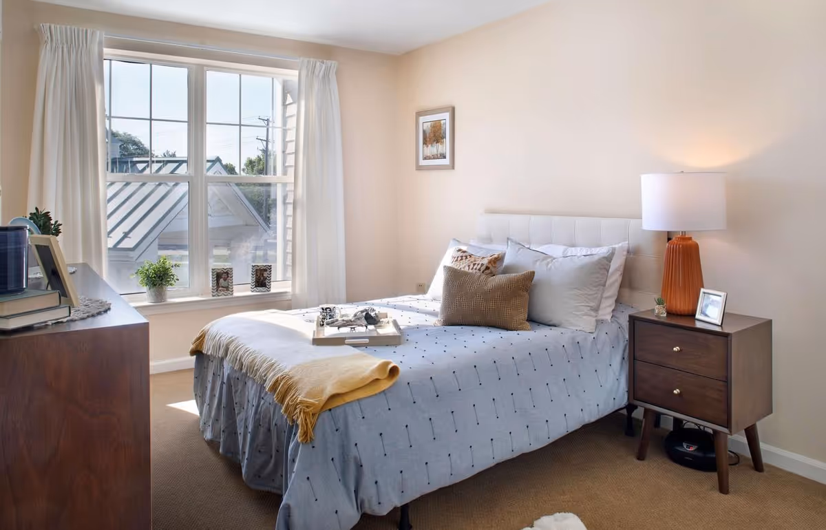 A bright and cozy bedroom with a large window letting in natural light. The bed is made with a light blue patterned comforter, several pillows, and a yellow throw blanket. There is a wooden nightstand with a lamp, a small plant, and a framed photo. A wooden dresser with books, a plant, and framed photos is positioned near the window. The walls are painted a soft beige color.
