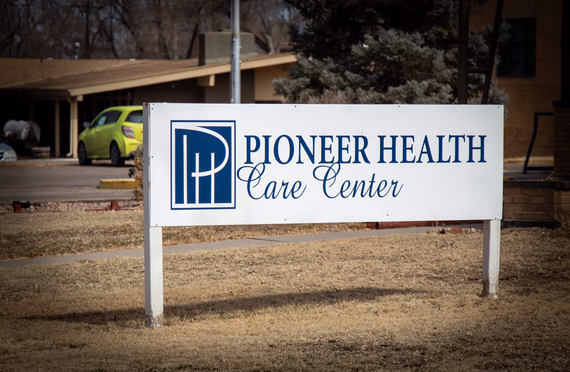 Outdoor view of a white sign with blue text that reads 'Pioneer Health Care Center' placed on a grassy area with a building and a green car in the background.