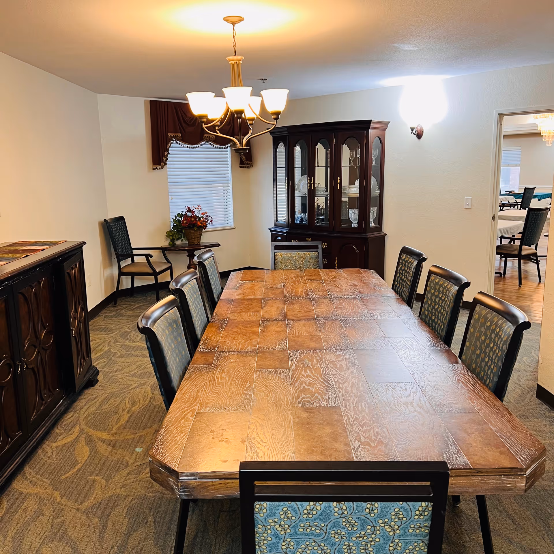 A dining room with a long wooden table surrounded by eight chairs with patterned upholstery. A chandelier with six lights hangs above the table. Against the wall is a dark wooden china cabinet displaying glassware and dishes. There is a small round table with a flower arrangement near a window with closed blinds and a valance. The room has carpeted flooring and light-colored walls. Through an open doorway, another room with tables and chairs is visible.