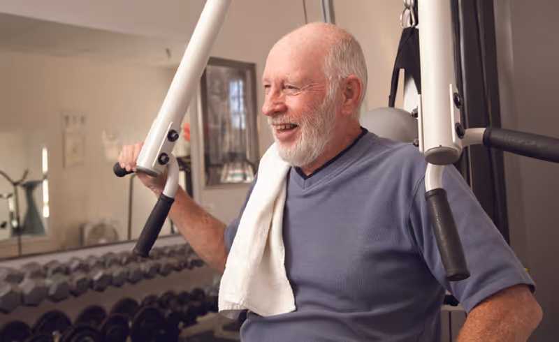 An elderly man with a white beard and short hair is smiling while using a weight machine in a gym. He is wearing a blue shirt and has a white towel draped over his shoulder. Behind him, there are dumbbells on a rack and a large mirror reflecting part of the room.