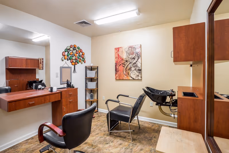 Interior view of a hair salon area in a senior living facility with two black salon chairs, a hair washing station with a black sink, wooden cabinets, a large mirror, and decorative wall art including a colorful tree sculpture and an abstract painting.