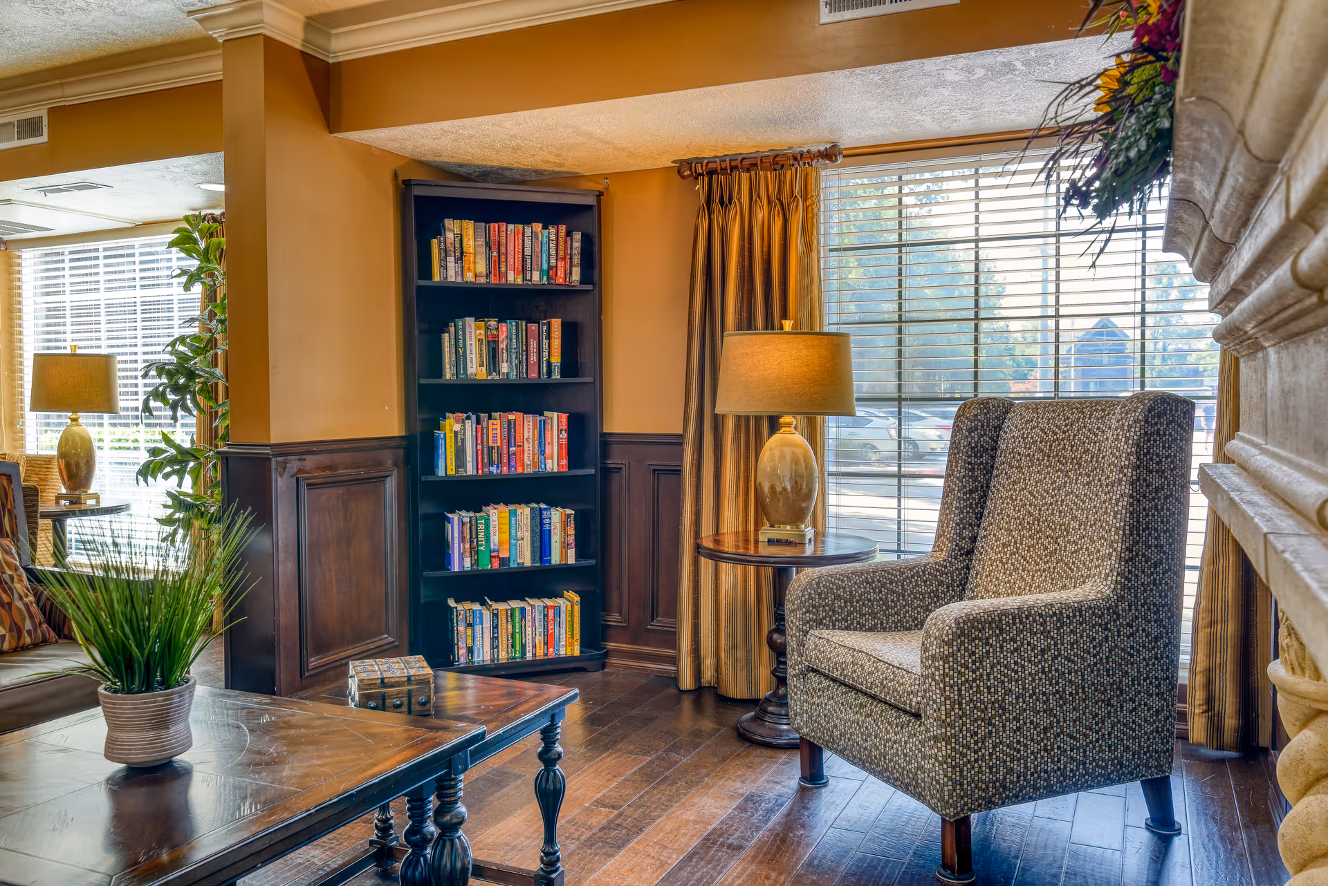 Cozy living room area with a patterned armchair next to a round wooden side table with a lamp. A tall black bookshelf filled with books is against the wall. Large windows with blinds and golden curtains let in natural light. A wooden coffee table with a potted plant and a decorative box is in the foreground. The room has warm tones with wood paneling and hardwood floors.