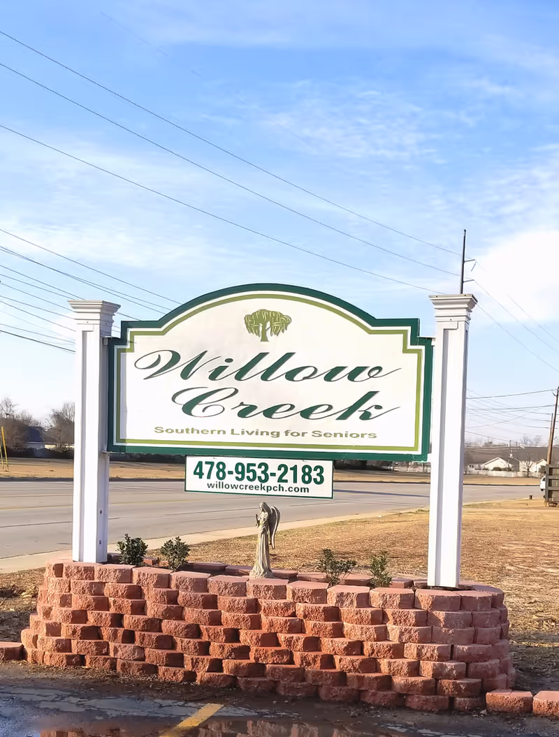 A freestanding roadside sign for Willow Creek assisted living with a phone number and a small brick planter below.