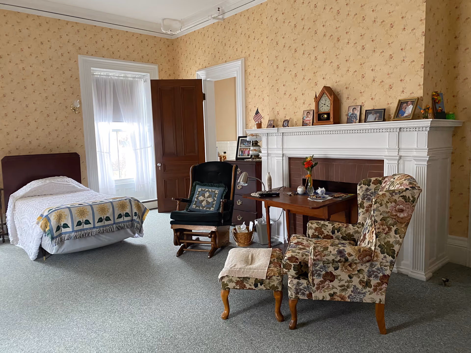 A bright resident bedroom with a single bed, a floral armchair and ottoman, a rocking chair, desk, and a white decorative fireplace mantel with framed photos.