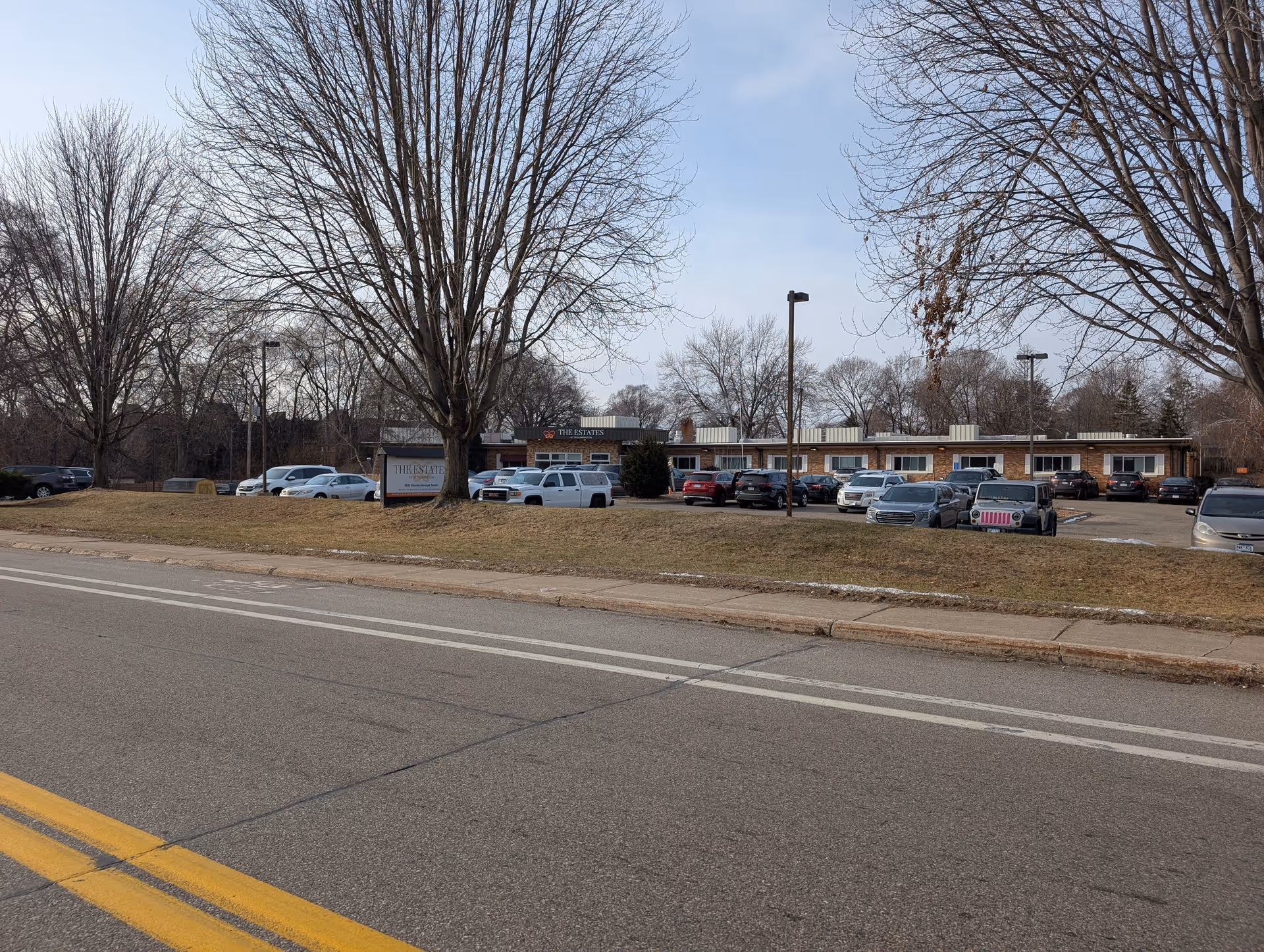 Exterior front view of The Estates at Bloomington building with parked cars, a lawn and bare trees along a street.