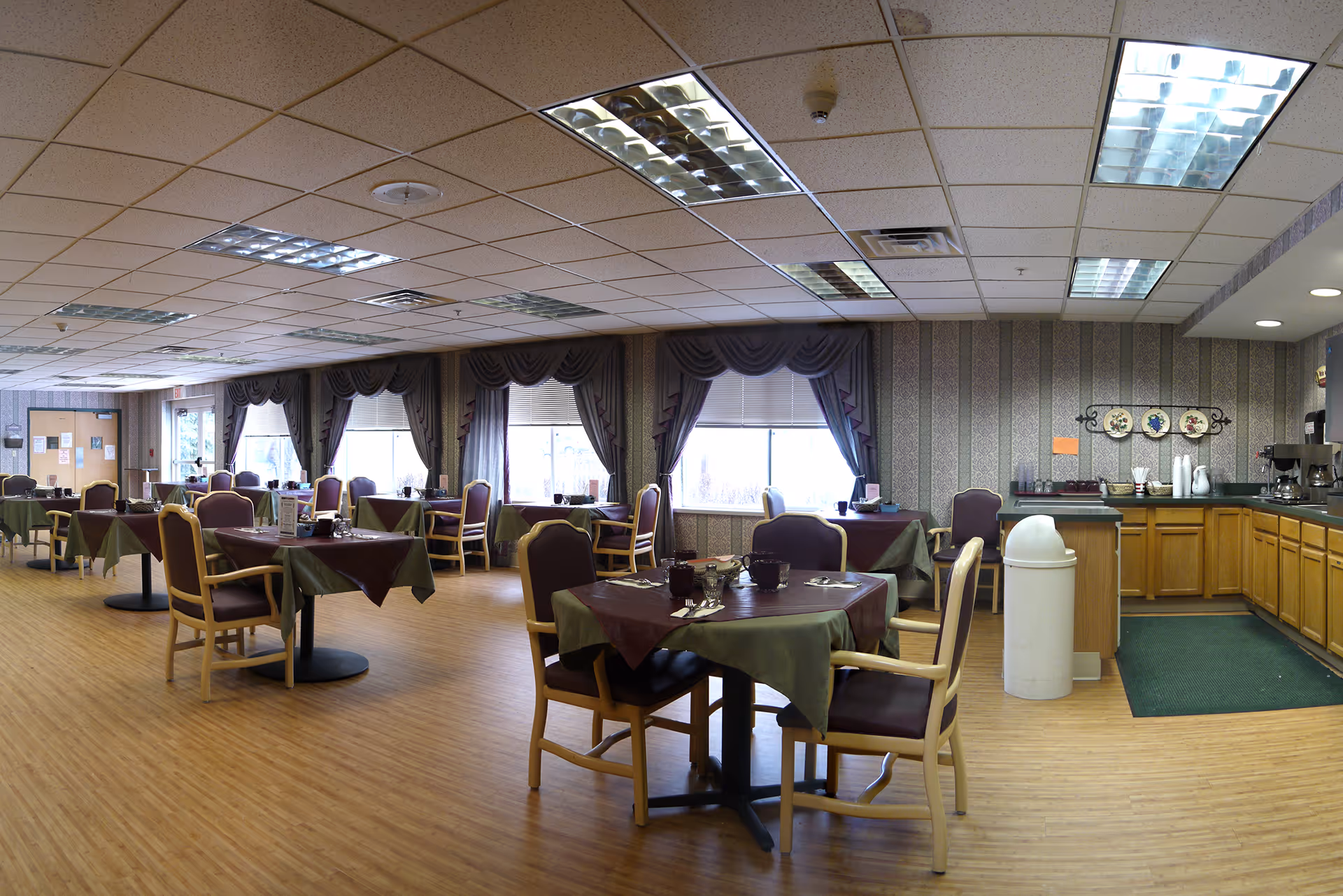 A spacious dining room in Pleasantview Care Center with multiple tables covered in green and brown tablecloths, each surrounded by wooden chairs with purple cushions. Large windows with decorative curtains let in natural light. To the right, there is a kitchenette area with wooden cabinets, a countertop, a coffee maker, and a white trash bin. The floor is wooden, and the ceiling has fluorescent lighting panels.