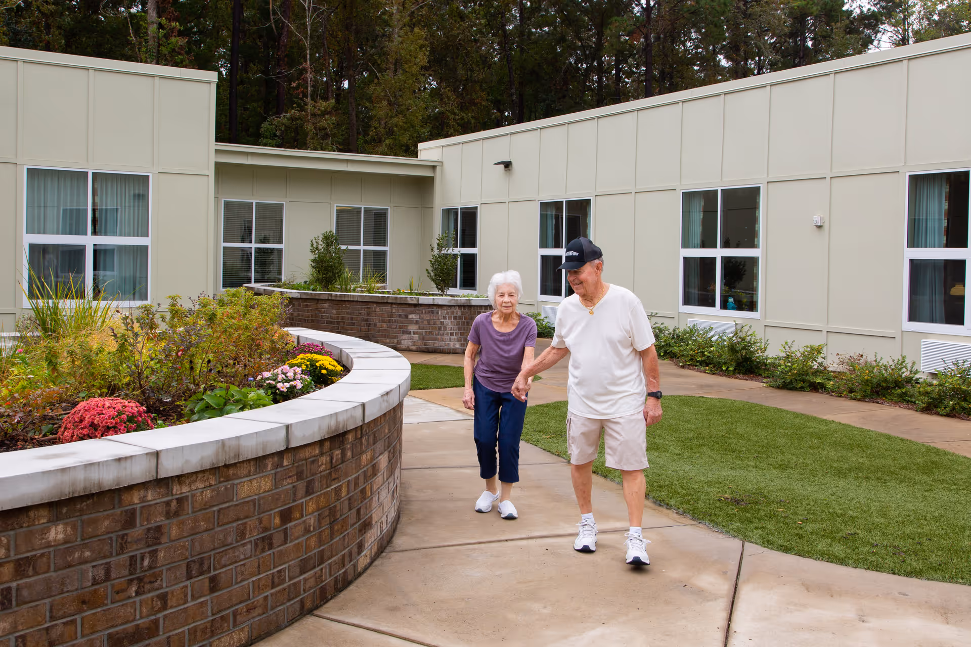 An elderly couple holding hands and walking along a curved concrete pathway in a garden courtyard area of a senior living facility. The courtyard features a raised brick planter with colorful flowers and greenery, surrounded by a building with large windows and beige paneling. Trees are visible in the background.