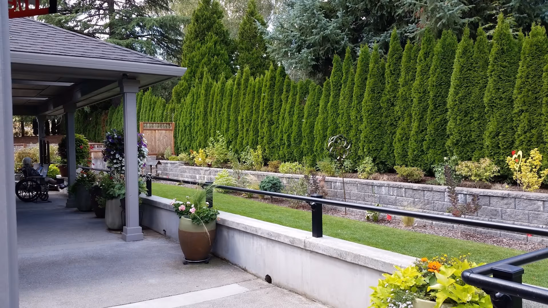 Outdoor patio area at a senior care home with a covered walkway, potted plants, a wheelchair, and a landscaped garden with tall green shrubs and flowers along a stone retaining wall.