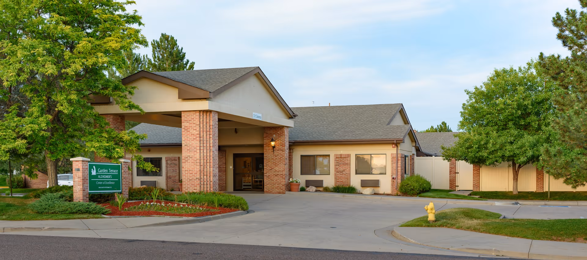 Exterior view of Garden Terrace at Aurora, a single-story building with brick pillars and a covered entrance. The surrounding area includes green trees, shrubs, and a neatly maintained lawn with a fire hydrant near the driveway.