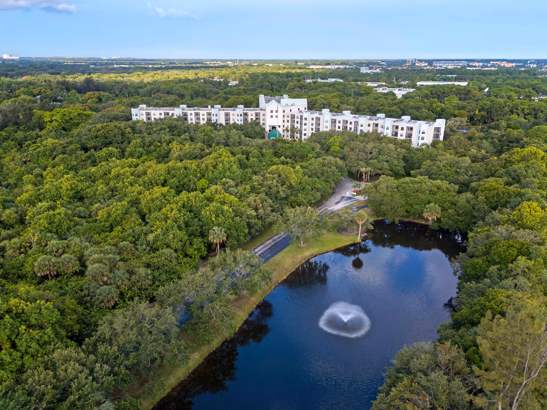 Aerial view of Brookdale Palm Beach Gardens senior living facility surrounded by dense green trees with a large pond featuring a water fountain in the foreground.