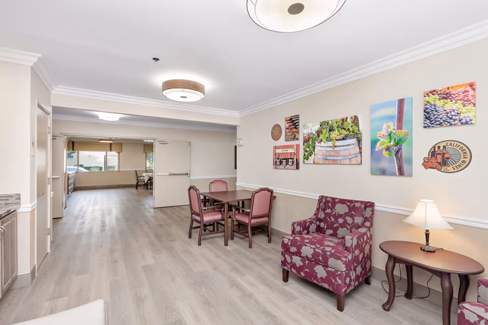 A bright interior common area in Brittany House Residential Memory Care featuring light wood flooring, a small square table with four pink cushioned chairs, a floral patterned armchair, a side table with a lamp, and colorful wall art depicting grapes, wine barrels, and vineyard scenes.