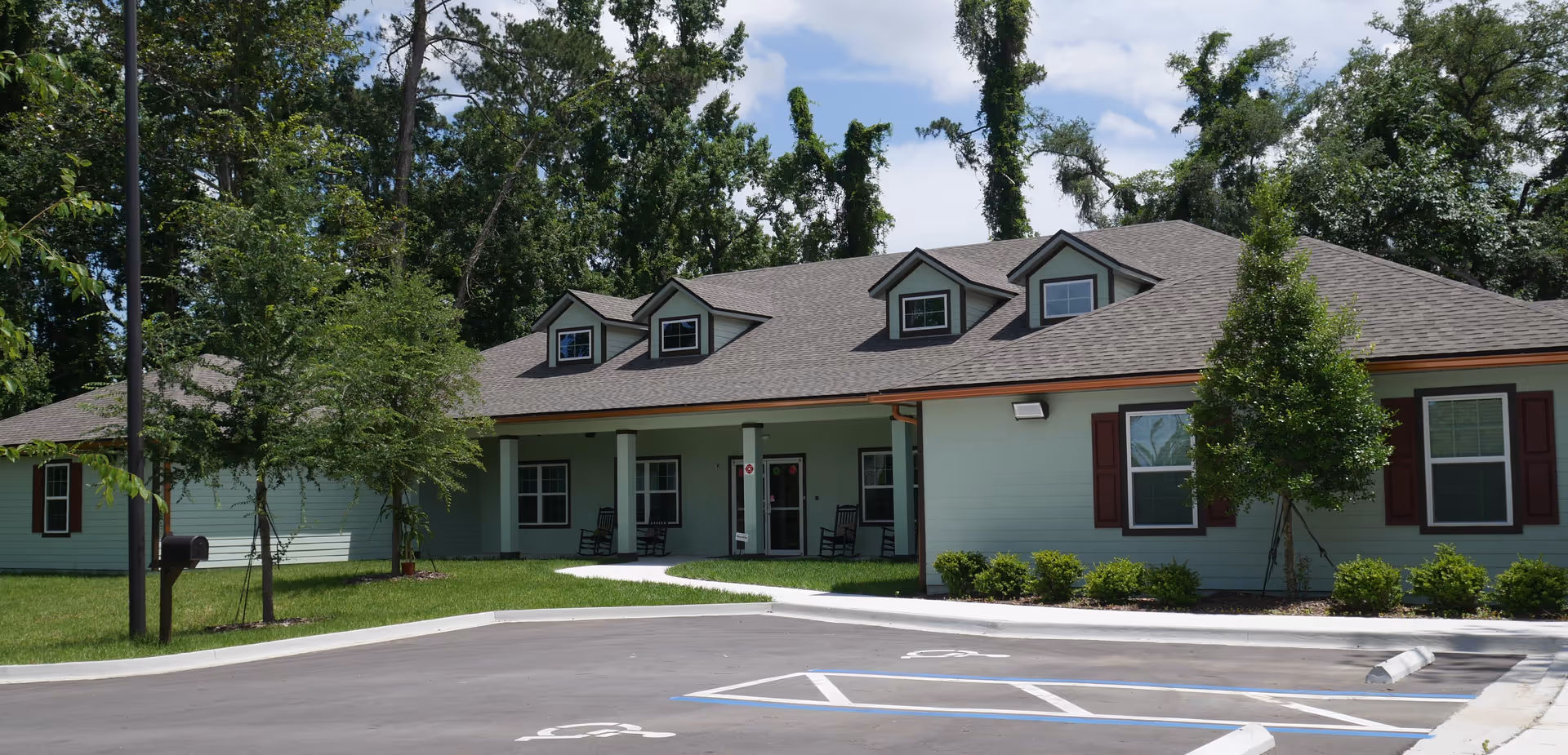 Single-story light-green building with a covered front porch, dormer windows, landscaping, and a parking lot with handicap spaces.