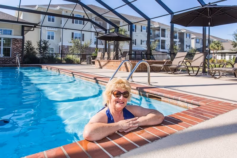 A smiling elderly woman wearing sunglasses and a blue swimsuit is relaxing in a swimming pool with a brick edge. The pool area is enclosed with a screened structure, and there are lounge chairs and umbrellas on the pool deck. In the background, there are multi-story residential buildings and some greenery.