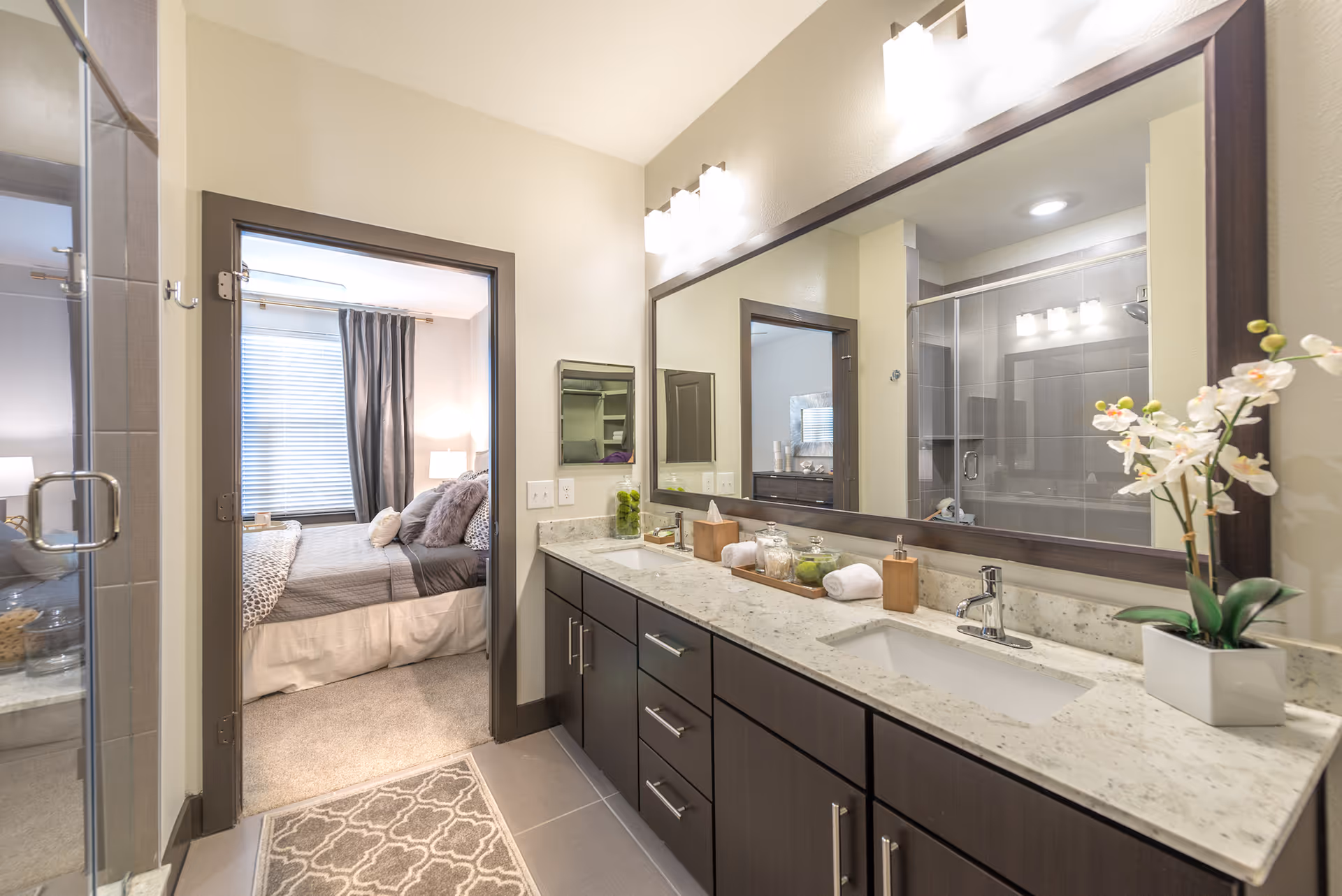 Modern bathroom with a large mirror above a double sink vanity with dark wood cabinets and a light-colored countertop. The bathroom has a glass-enclosed shower on the left and a doorway leading to a bedroom with a bed, pillows, and curtains visible.