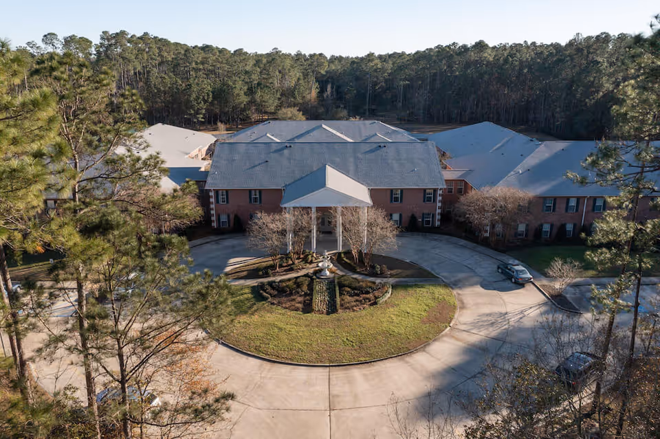 Aerial view of a large brick building with a gray roof surrounded by trees. The building has a circular driveway with a landscaped island in the center featuring a fountain. Several cars are parked near the building, which is set in a wooded area.
