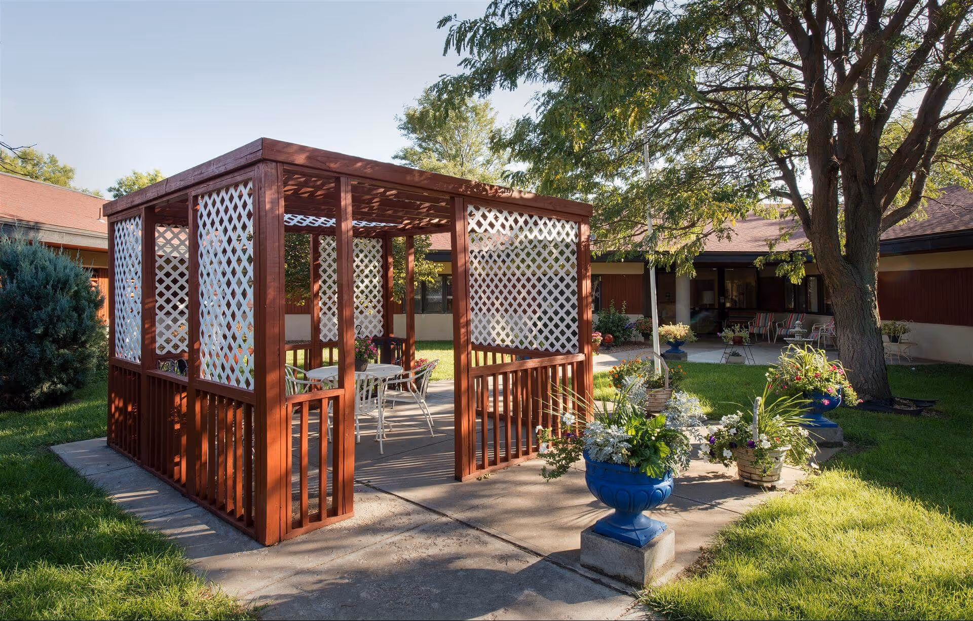 Wooden outdoor gazebo with white lattice panels and seating in a grassy courtyard with potted flowers and trees in front of a low red-roofed building.