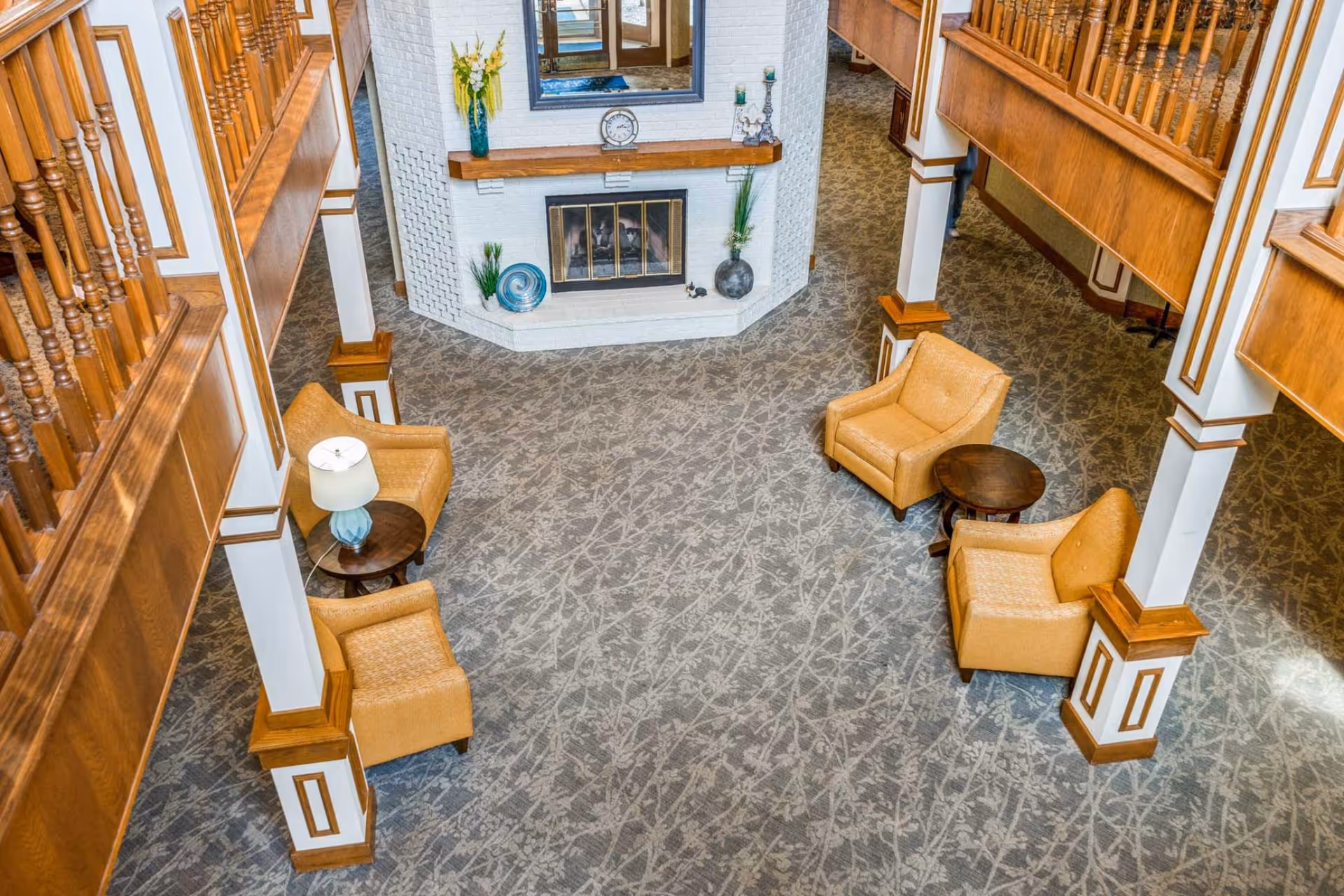 View from above of a spacious living room area with patterned carpet, four beige armchairs arranged around two small round wooden tables, a white brick fireplace with decorative items on the mantel, and wooden railings on the upper level.