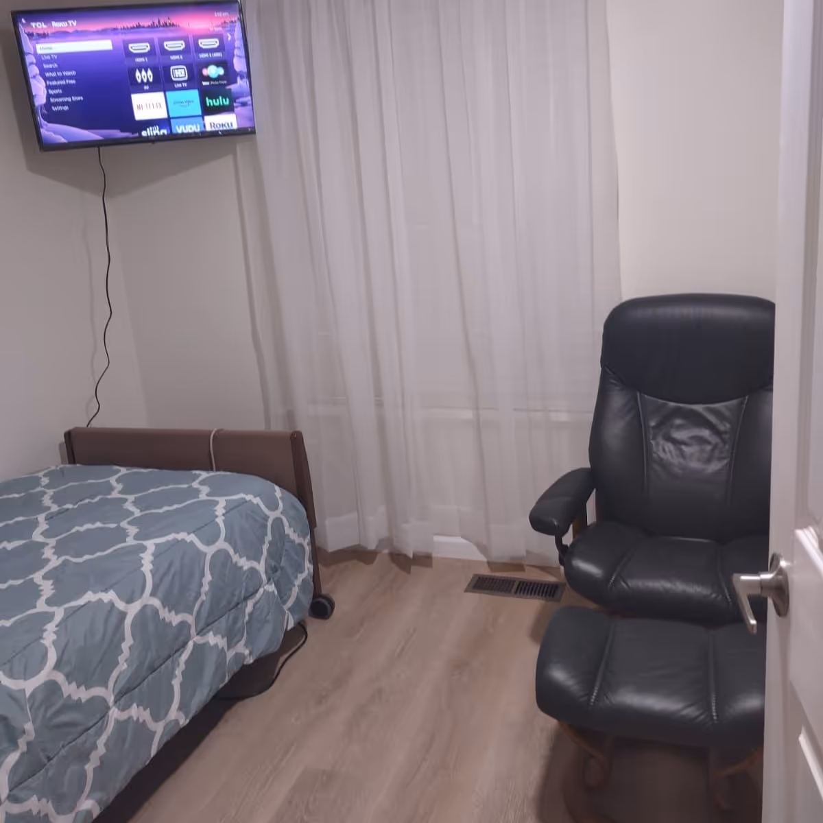 Small bedroom with a single bed, wall-mounted TV, and a black leather chair by a window with sheer curtains.
