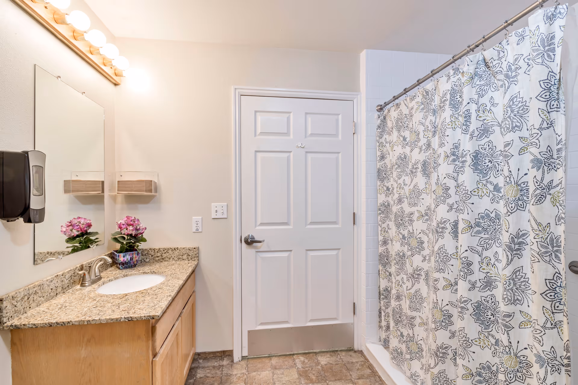 Well-lit bathroom with a granite vanity and sink on the left, a closed door in the center, and a floral shower curtain on the right.