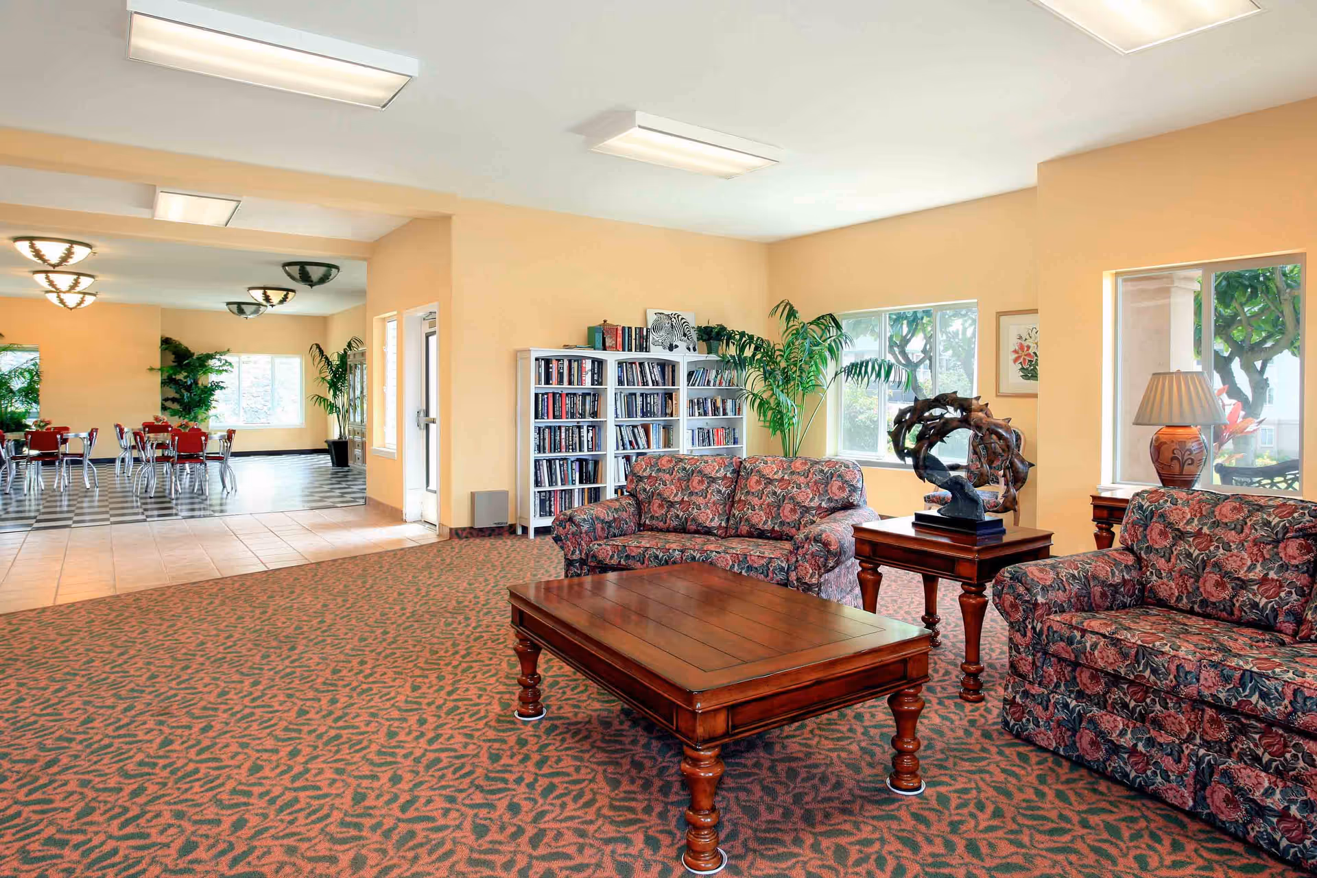Bright communal living room with floral sofas, a wooden coffee table, bookshelves and an adjacent dining area visible in the background.