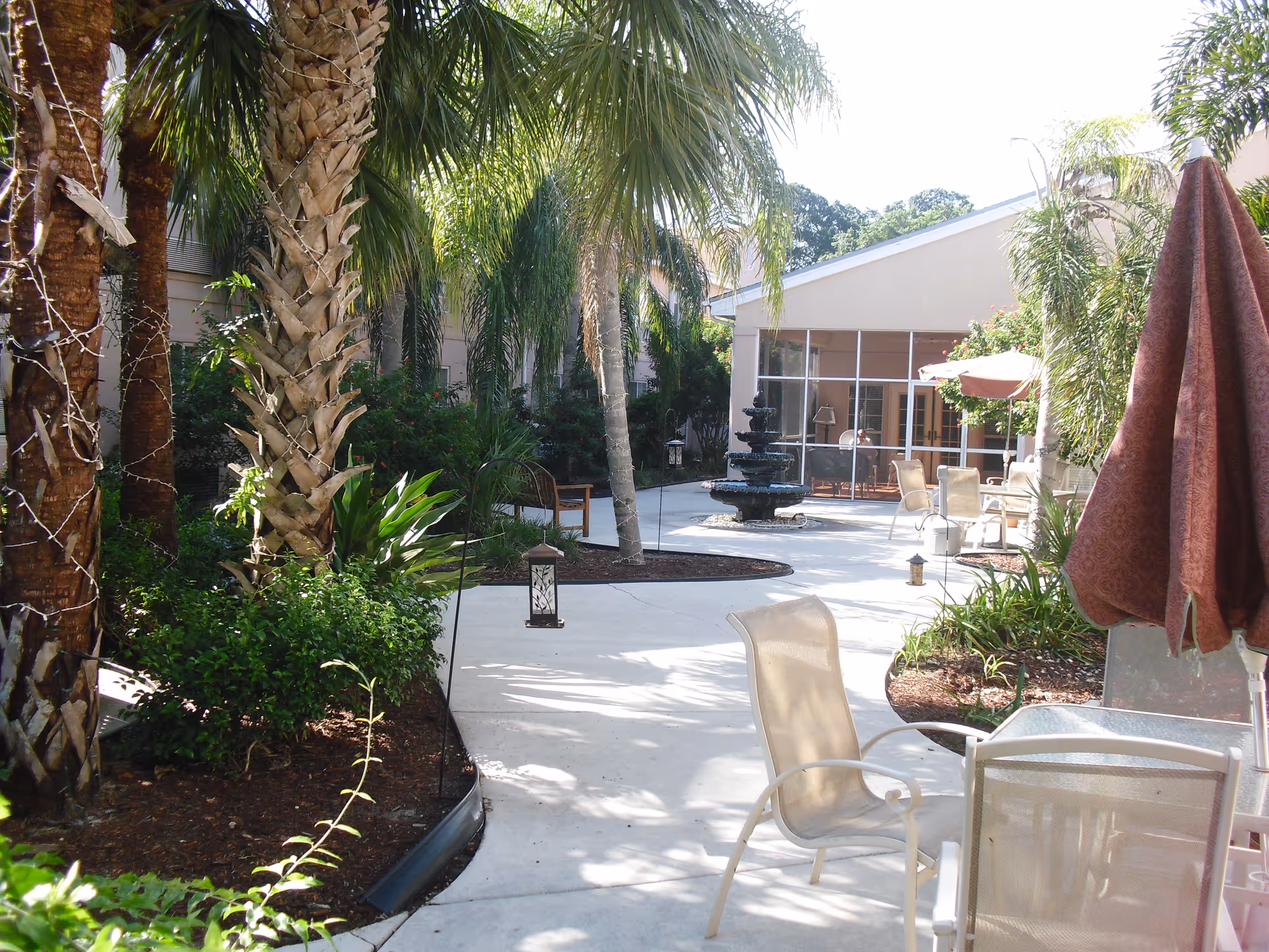Outdoor courtyard area with palm trees, plants, and a concrete pathway. There are several chairs and tables with umbrellas, and a multi-tiered black fountain near a building with large windows.