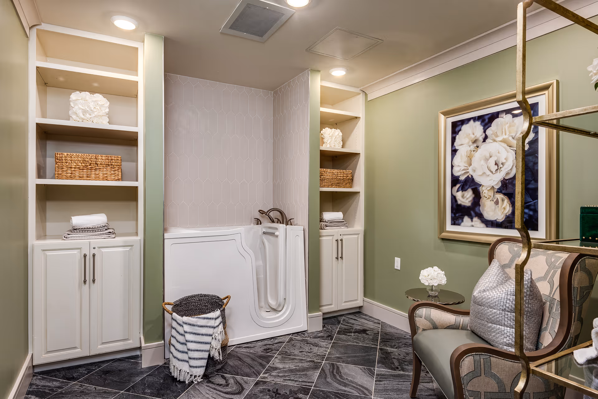 A well-lit bathroom featuring a walk-in bathtub with a door, flanked by built-in white shelves and cabinets on both sides. The shelves hold decorative items including folded towels, a wicker basket, and white coral-like decor. The walls are painted light green, and there is a framed floral artwork hanging on the right wall. A comfortable armchair with a patterned cushion and a small round side table with a white flower arrangement are also visible. The floor is covered with dark, marbled tiles.