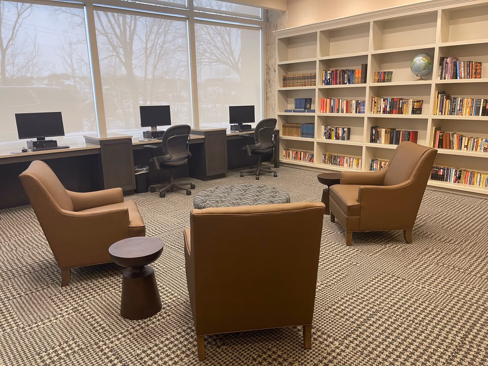 A cozy room with three brown armchairs arranged around a patterned ottoman in the center. Behind the chairs, there are three computer workstations with black office chairs and monitors. A large bookshelf filled with books and a globe is against the wall. Large windows with roller shades let in natural light.