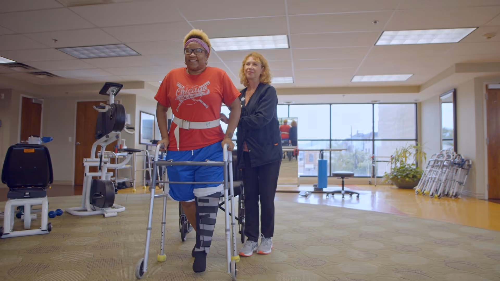 A woman using a walker with a leg brace is assisted by a caregiver in a spacious rehabilitation or physical therapy room with exercise equipment and large windows in the background.