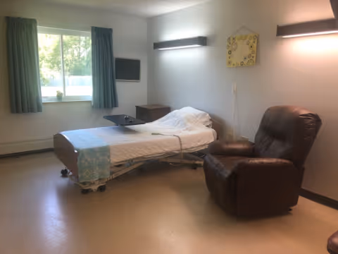 A simple, clean room with a hospital-style bed covered with white sheets and a light blue blanket at the foot. Next to the bed is a dark brown recliner chair. The room has a window with blue curtains, a small wooden nightstand, a wall-mounted TV, and two modern wall lights. The walls are light-colored and the floor is a neutral tone.