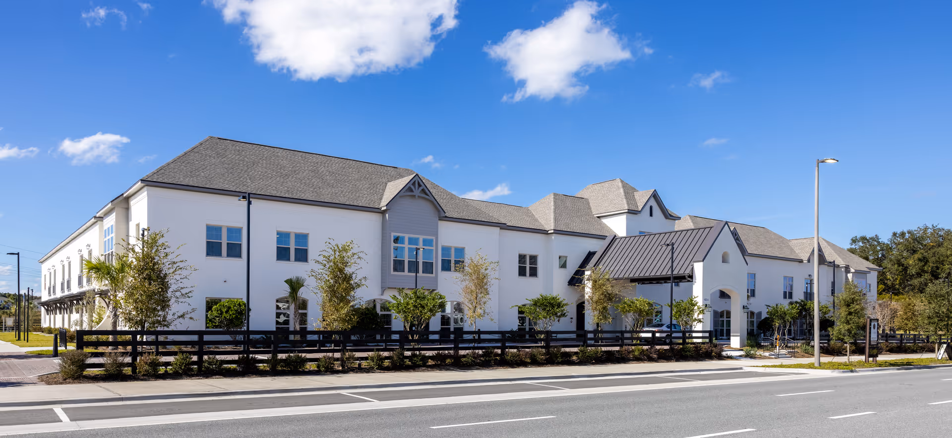Exterior view of The Blake at Viera, a large two-story senior living facility with white walls, multiple windows, a gray roof, and a black fence surrounding landscaped greenery. The building is situated along a road under a clear blue sky.