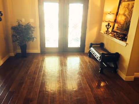 Entryway with double glass doors featuring decorative wrought iron designs, a dark wooden bench with a cushion on the right, a small lamp and framed artwork on a shelf above the bench, and a potted plant on the left side. The floor is made of polished dark wood.