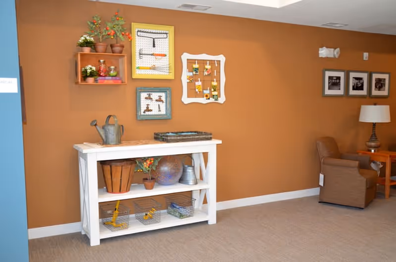 Interior view of a room with a brown wall featuring decorative framed items and small potted plants. A white wooden table holds various decorative objects including a watering can, a basket, and a metal pitcher. To the right, there is a brown upholstered armchair next to a wooden side table with a lamp and framed pictures on the wall above.