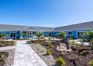 Landscaped courtyard with a paved walkway leading to a single-story blue building under a clear blue sky.
