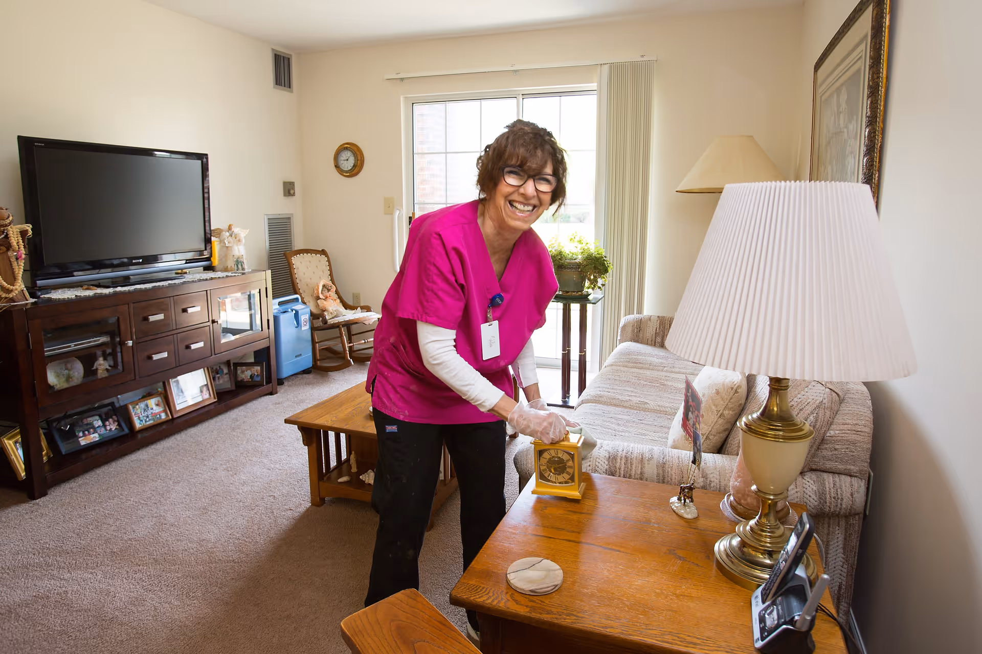 A smiling woman in a magenta scrub top and glasses is cleaning a small clock on a wooden side table in a cozy living room. The room features a beige couch, a wooden coffee table, a large flat-screen TV on a wooden cabinet, a lamp with a pleated shade, and a sliding glass door letting in natural light.