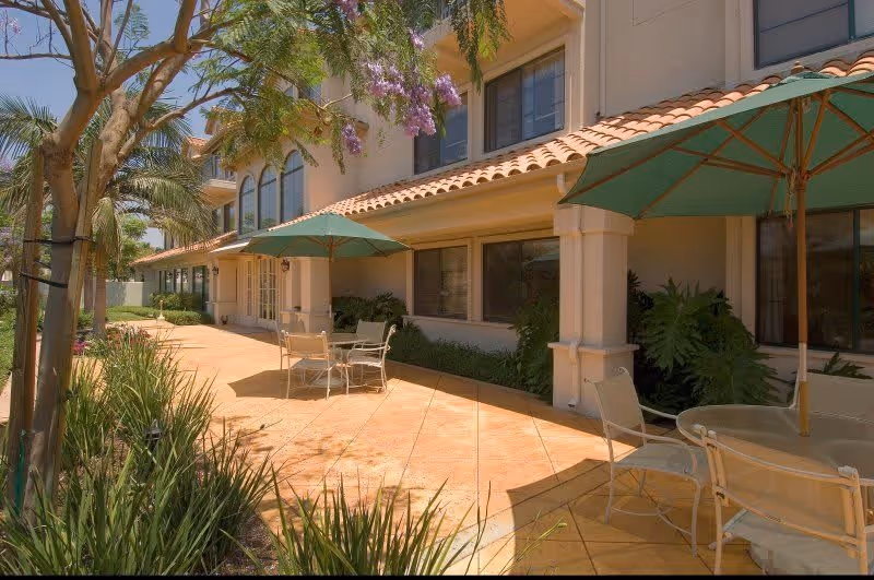 Outdoor patio area at Aegis of Granada Hills with tiled flooring, several tables with green umbrellas, and chairs. The patio is adjacent to a beige building with large windows and surrounded by greenery including palm trees and flowering plants.