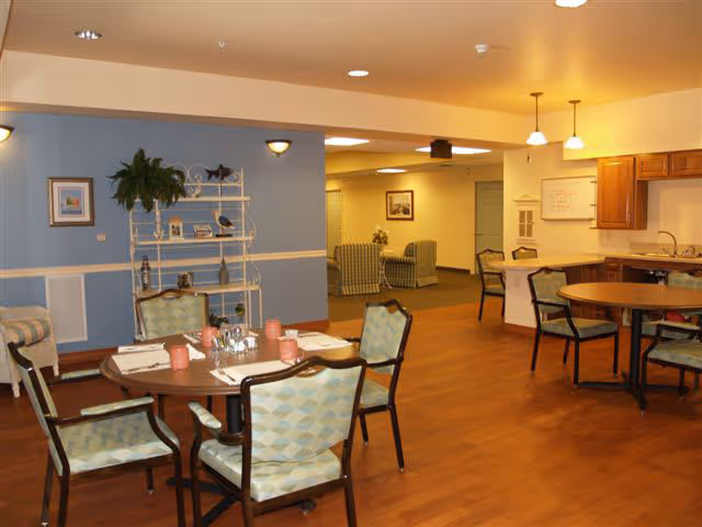 A dining area in an assisted living facility with round tables set with cups and napkins, cushioned chairs, a kitchen area with wooden cabinets and a sink, and a seating area with armchairs in the background. The room has warm lighting and wooden flooring.