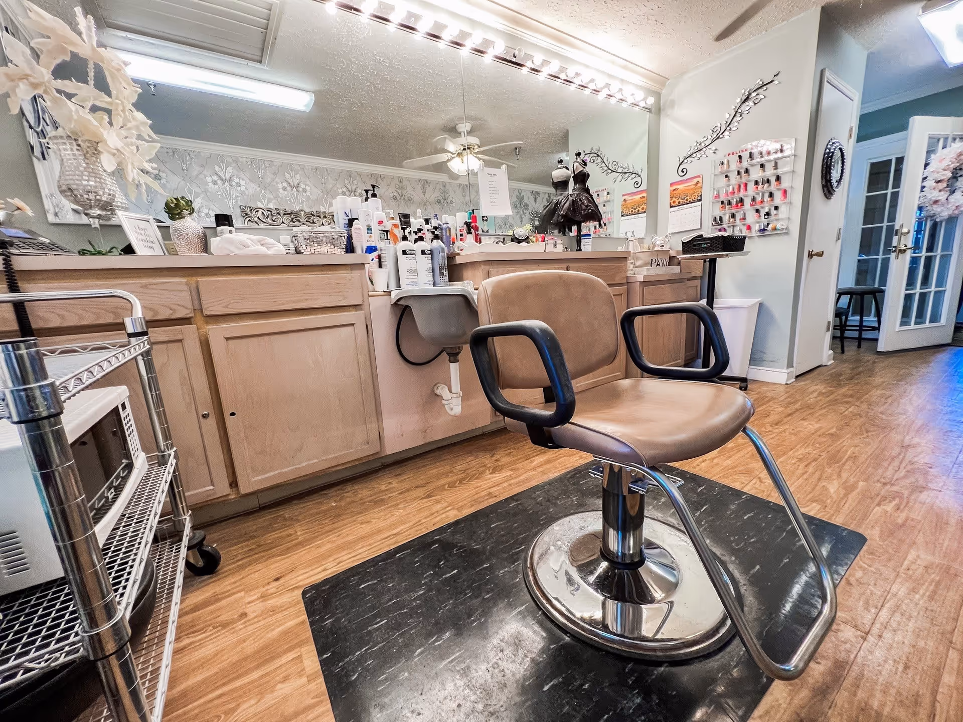 Interior view of a salon area with a brown salon chair on a black mat in front of a counter filled with various hair and beauty products. A large mirror with lights above it reflects the room, which has wooden flooring and light-colored walls. There is a metal cart with a microwave on the left side and a door with glass panels in the background.