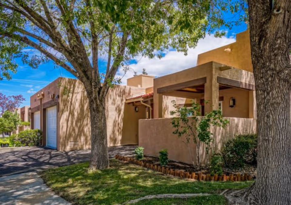 Exterior view of a southwestern-style building with tan stucco walls, a covered patio, and a garage door. The scene includes green grass, trees with leafy branches, and a bright blue sky with some clouds.