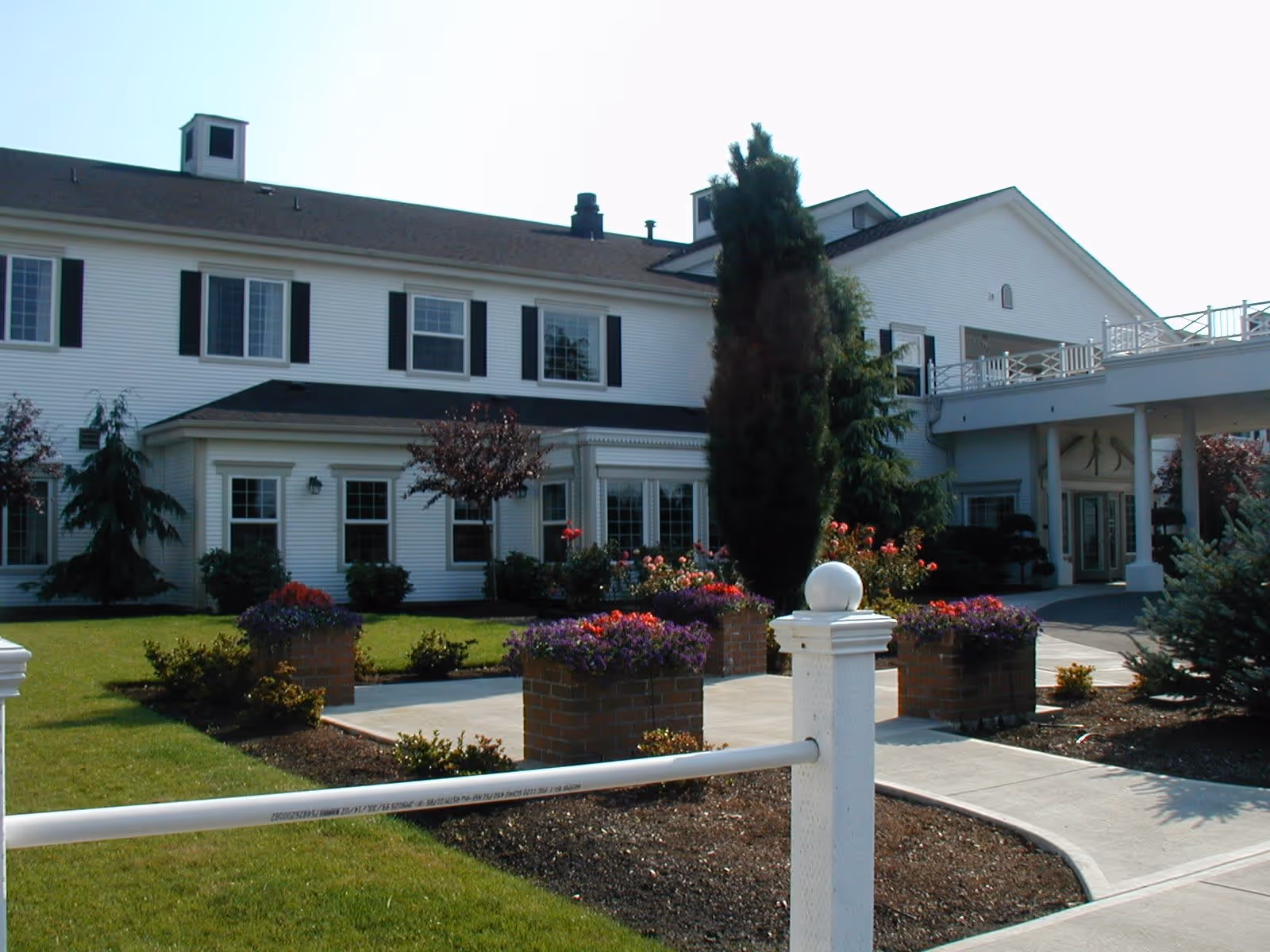Exterior view of a two-story white building with black shutters, surrounded by a well-maintained garden with colorful flowers and shrubs. A paved walkway leads to the entrance, which features a covered porch supported by columns.