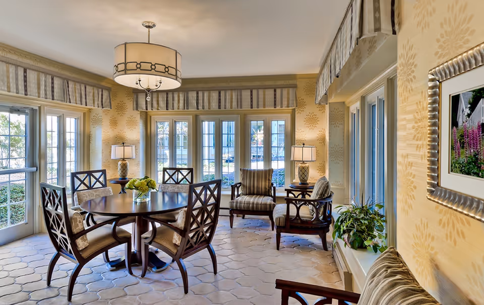 Bright, elegantly furnished common room with a round dining table surrounded by chairs, armchairs, lamps, and large windows.