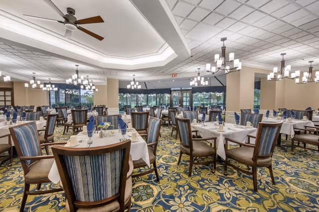 Spacious dining room with multiple tables covered in white tablecloths, each set with blue napkins, plates, and silverware. The room features patterned carpet, large windows allowing natural light, and modern chandeliers hanging from the ceiling.