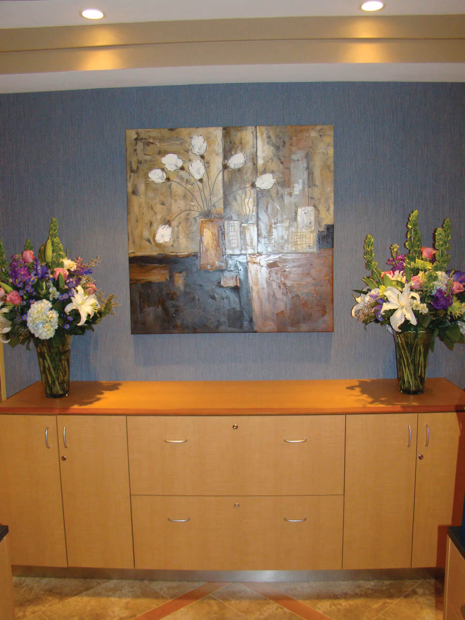A wooden cabinet with two large floral arrangements in glass vases on each end, placed against a blue textured wall. Above the cabinet hangs a large abstract painting featuring white flowers in vases with a mix of earthy tones.
